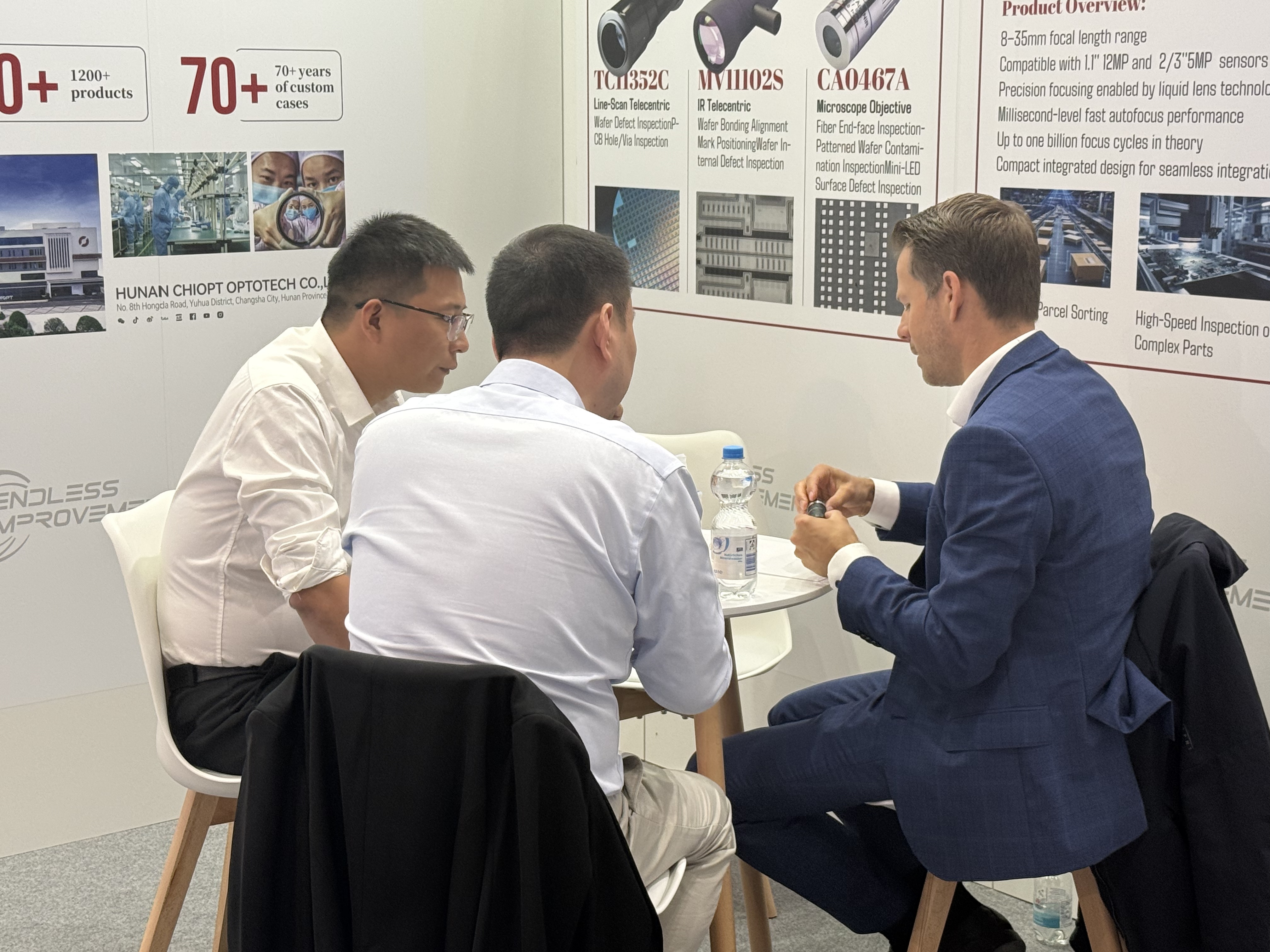 Engineers discussing a lens at the CHIOPT booth in front of the semiconductor inspection lens display wall